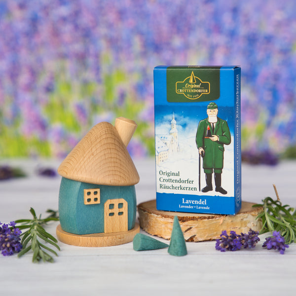 Ein Holzwaren Egermann Räucherhäuschen rund aus Holz blau/natur und eine Schachtel Lavendel-Räucherkegel stehen auf einem Tisch inmitten von Lavendelblüten, im Hintergrund ein verschwommenes Feld. Die Erzgebirgische Handarbeitsbox zeigt eine Figur in traditioneller Kleidung.