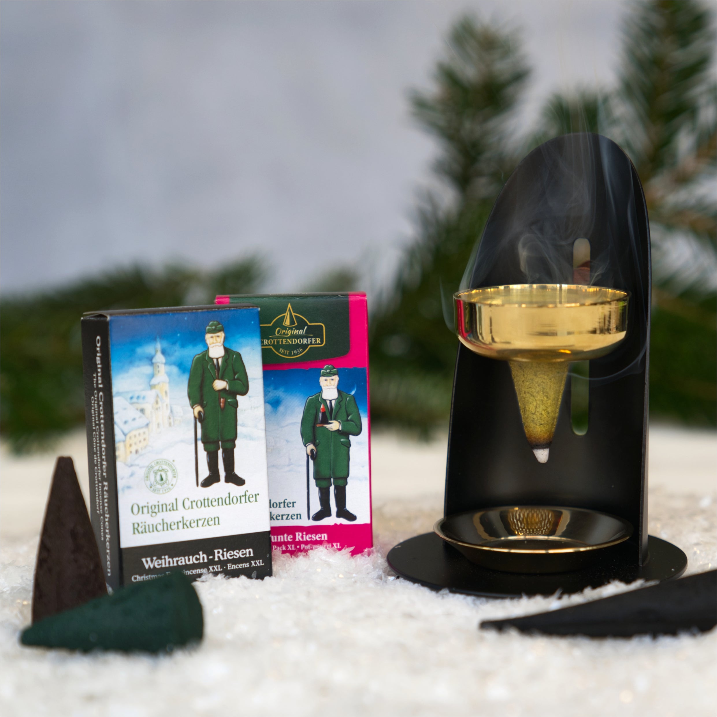 Eine Winterlandschaft zeigt zwei Räuchergefäße mit einer traditionellen Figur und deutschem Text. Im Vordergrund ist der höhenverstellbare Weihrauchbrenner Messing/Schwarz von Original Crottendorfer zu sehen, der inmitten von Schnee und Tannenzweigen einen brennenden Räucherkegel hält und so exquisite Handarbeit hervorhebt.