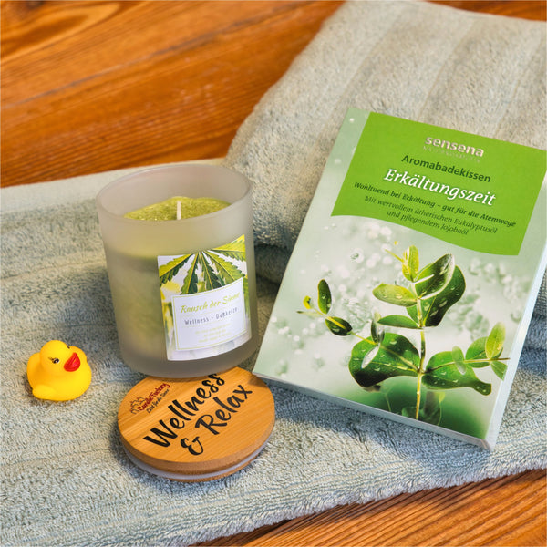 Eine Wellness-Duftkerze "Rausch der Sinne" von Candle Factory, eine Dose Erkältungszeit-Aromabadesalz, ein hölzerner Wellness & Relax-Untersetzer, eine gelbe Gummi-Ente und ein gefaltetes grünes Handtuch sind auf einer Holzfläche angeordnet.