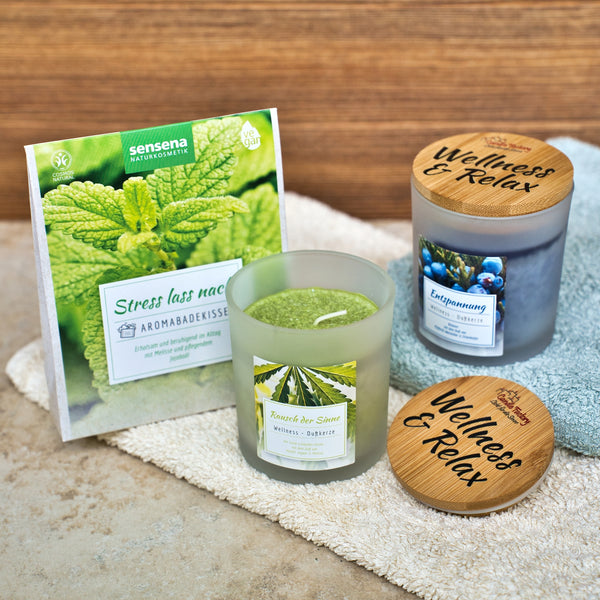 Ein grünes Kräutersäckchen und zwei Candle Factory Wellnessduftkerzen "Rausch der Sinne" - grüne und blaue Stearin-Kerzen mit gravierten Holzdeckeln - auf Holz, ideal für Ihre Naturkosmetik-Routine.