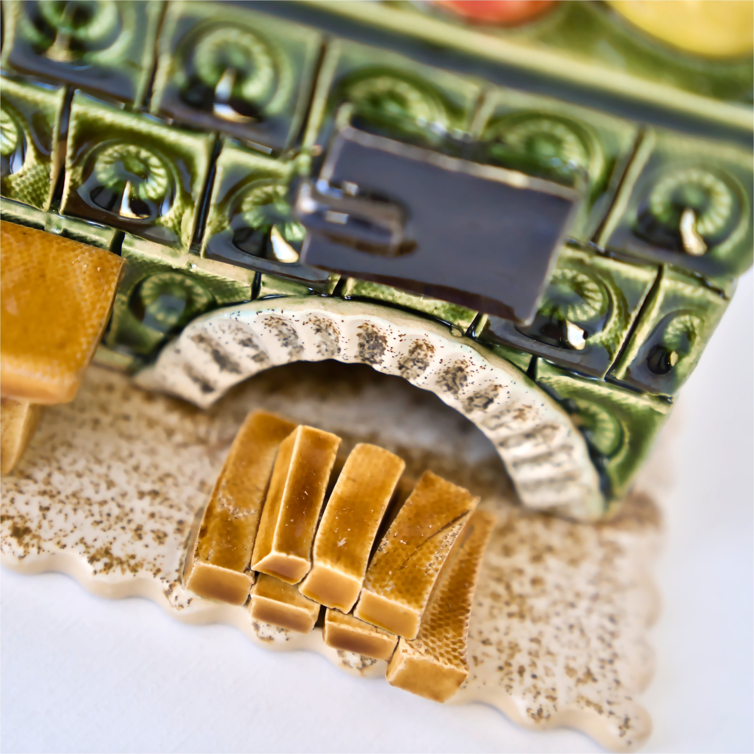 Nahaufnahme der Miniatur „Keramik-Küchenherd Zuckerbäckerei grün, eckig“ von der Keramikwerkstatt Carola Busch, die einem alten Kachelofen mit grünen Kacheln, einer schwarzen Tür und Holzscheiten unter einem weißen Bogen ähnelt. Er ruht auf einer leicht strukturierten Oberfläche.