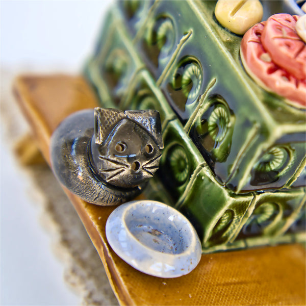 Eine kleine graue Katzenfigur sitzt neben einem grünen, mit Mustern verzierten „Keramik-Küchenherd Zuckerbäckerei“ der Keramikwerkstatt Carola Busch. Davor steht eine blaue Untertasse auf einem goldenen Kissen mit Spitzenrändern, die an erzgebirgisches Kunsthandwerk erinnern.