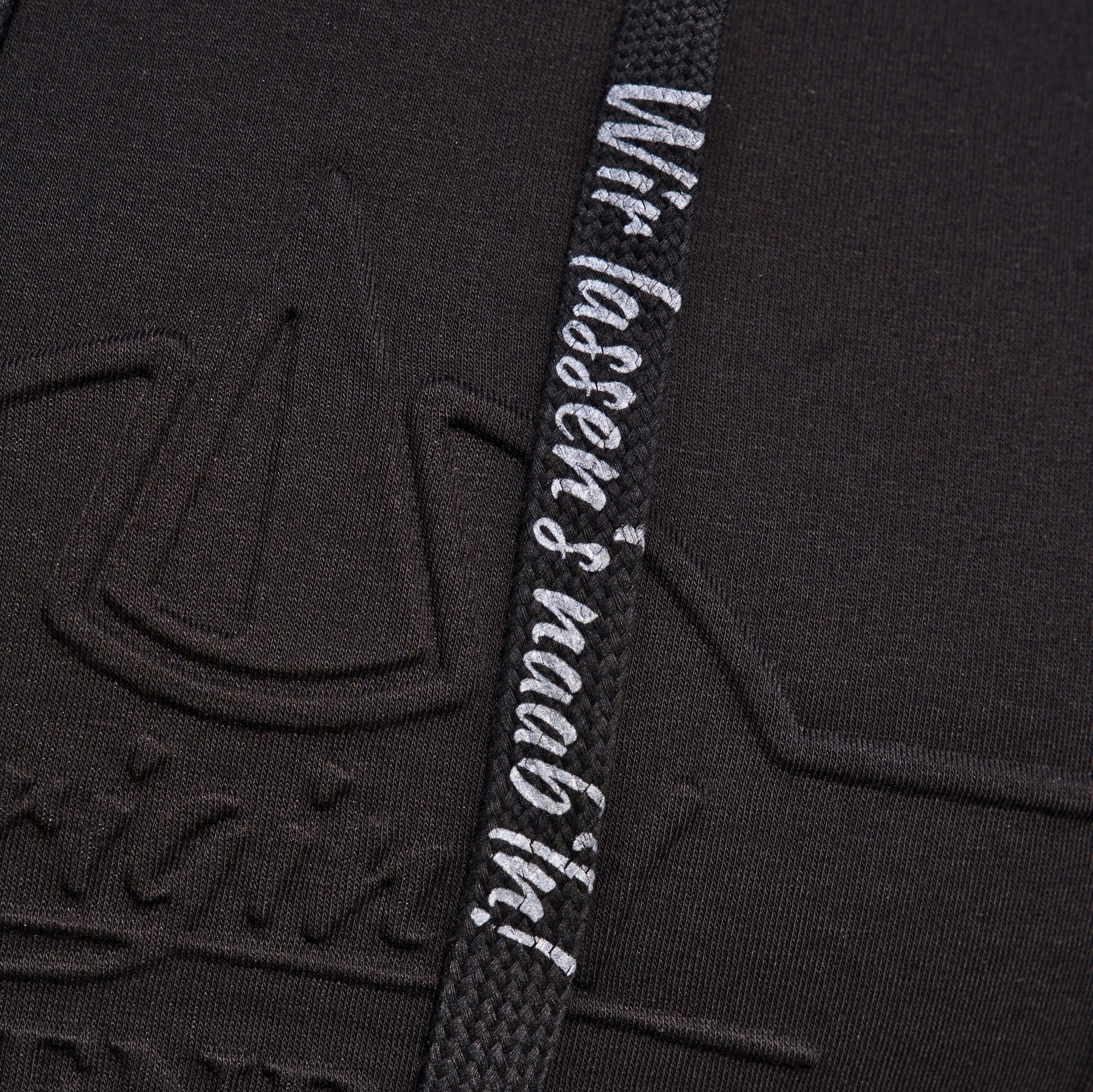 Eine Nahaufnahme des schwarzen strukturierten Stoffes des Original Crottendorfer Kapuzenpullovers zeigt ein geprägtes Logo und einen Riemen mit weißem deutschem Text: „Wir lassens kraabbln!“. Ideal für einen Streetwear-Look, deutet der strapazierfähige Stoff Robustheit an und gibt gleichzeitig teilweise den Blick auf das Logo-Design frei.
