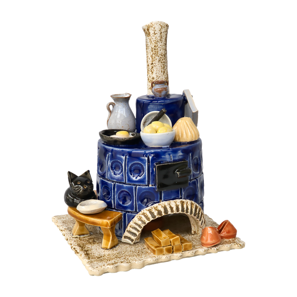 Der Keramik-Räucherofen Herd blau, rund der Keramikwerkstatt Carola Busch ist eine Keramikskulptur, die einen blauen Holzofen mit Schornstein darstellt, komplett mit Küchenutensilien, Brennholz, orangefarbenen Hausschuhen für gemütliche Wärme und einer schwarzen Katze auf einer Bank.