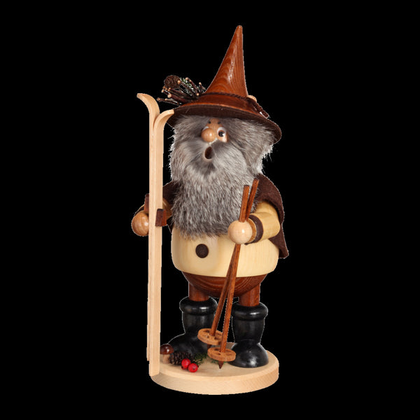 Der Wichtel Skifahrer von DWU Drechselwerkstatt Uhlig ist eine Räuchermann-Figur aus Holz, die einen bärtigen Mann mit Skiern und Stöcken darstellt, der einen spitzen Hut und einen Umhang trägt. Er steht auf einem runden Sockel mit Tannenzapfen und Beeren und zeigt den traditionellen Erzgebirge-Stil mit detaillierten Merkmalen.