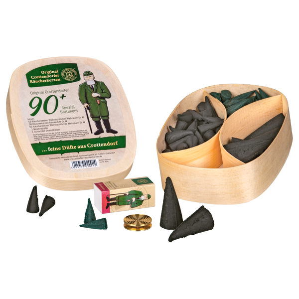 Das Geschenkset Spanschachtel 90+ von Original Crottendorfer Räucherkerzen enthält eine Holzkiste mit Fächern für schwarze Räucherkegel, einen Messinghalter, Streichhölzer und eine Verpackung mit einem Cartoon-Mann in grüner Kleidung.