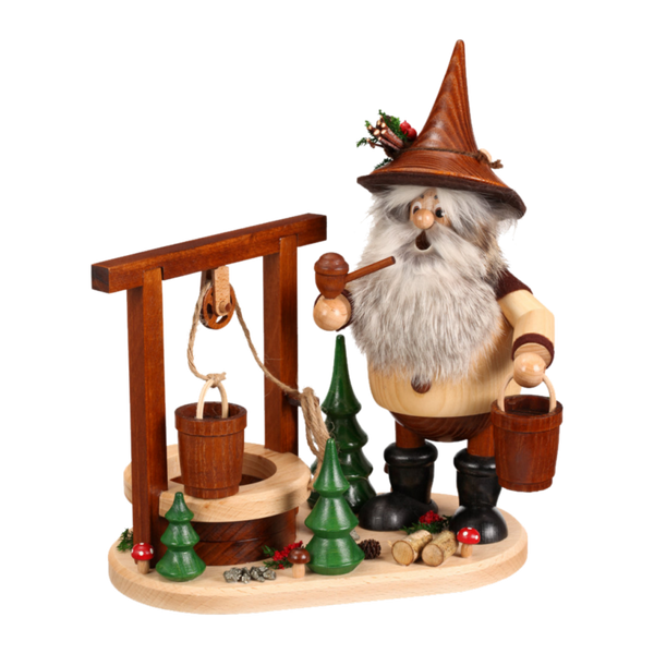 Das Erzwichtel am Brunnen von DWU Drechselwerkstatt Uhlig zeigt einen hölzernen Räuchermann-Zwerg mit Pfeife an einem Brunnen, der einen Eimer hält, inmitten von Mini-Weihnachtsbäumen und Baumstämmen und schafft so eine festliche Szene der erzgebirgischen Volkskunst.