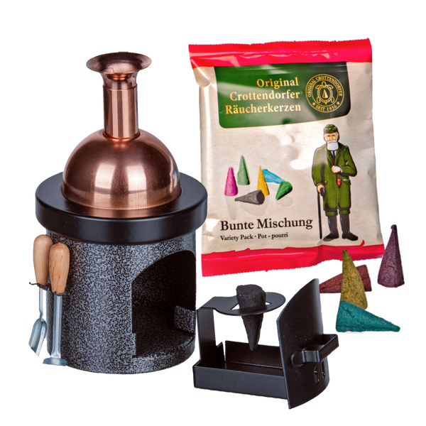 Ein kupfer- und schwarzfarbener Hartmut Richter Räucherofen-Braukessel in Form eines Kamins steht neben einer Packung verschiedener Räucherkegel mit bunten Kegelbildern und einer Figur in einem grünen Mantel. Zu sehen sind auch lose Kegel, ein Aschekasten und ein kleines Werkzeugset.