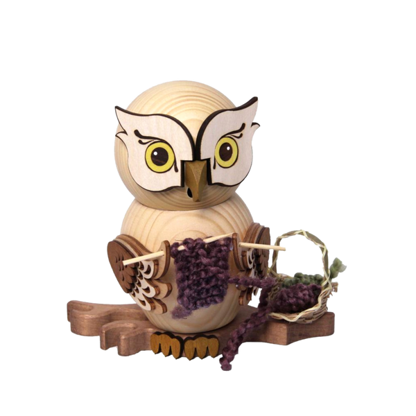 Die Rauchfigur Eule mit Strickzeug von Drechslerei Kuhnert zeigt eine Holzeule mit großen, ausdrucksstarken Augen, die einen lila-grünen Schal auf einem Ast strickt. Ihre detailliert geschnitzten Flügel und die glatte Oberfläche erinnern an Raucheulen.