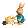Der Stupsi Hase mit Schubkarre von Drechslerei Kuhnert ist ein bezaubernder Holzhase mit großen Ohren, blauem Oberkörper und orangefarbener Fliege. Er schiebt eine Schubkarre mit einem gelben Küken und Stroh und hat ein schlicht bemaltes Gesicht, das die entzückende Essenz des Osterhasens einfängt.