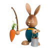 Der Stupsi Hase Möhrenangler von Drechslerei Kuhnert ist eine handgefertigte Hasenfigur aus Holz mit großen Ohren und einem aufgemalten Lächeln, die eine Angelrute mit einer Karotte hält. Sie trägt ein blaues Outfit und trägt einen Eimer mit kleinen Karotten.