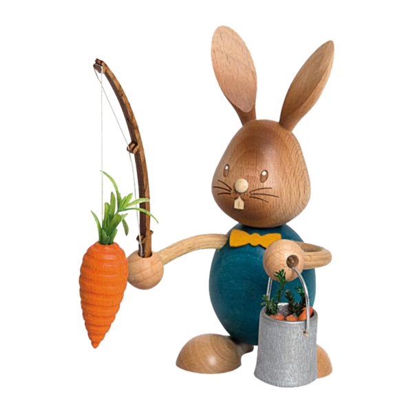 Der Stupsi Hase Möhrenangler von Drechslerei Kuhnert ist eine handgefertigte Hasenfigur aus Holz mit großen Ohren und einem aufgemalten Lächeln, die eine Angelrute mit einer Karotte hält. Sie trägt ein blaues Outfit und trägt einen Eimer mit kleinen Karotten.