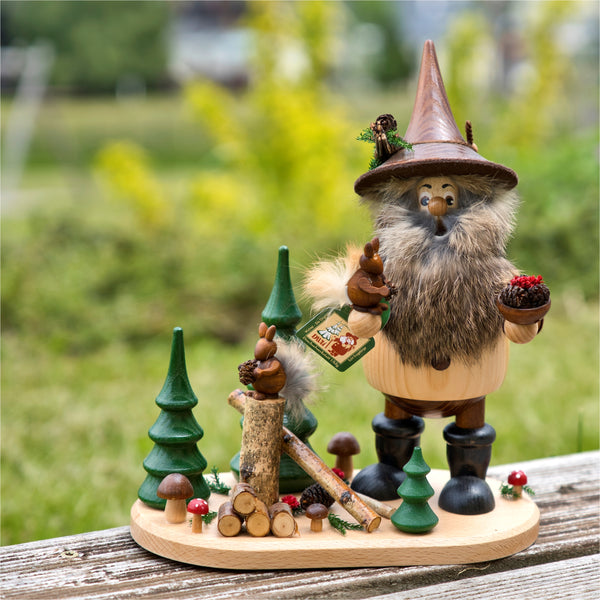 Das Waldwichtel mit Eichhörnchen von DWU Drechselwerkstatt Uhlig zeigt einen bärtigen Räuchermann mit spitzem Hut, der auf einem Holzsockel zwischen Bäumen, Baumstämmen und Pilzen ein Eichhörnchen und einen Korb hält. Diese erzgebirgische Kreation sticht vor der verschwommenen grünen Gartenkulisse hervor.