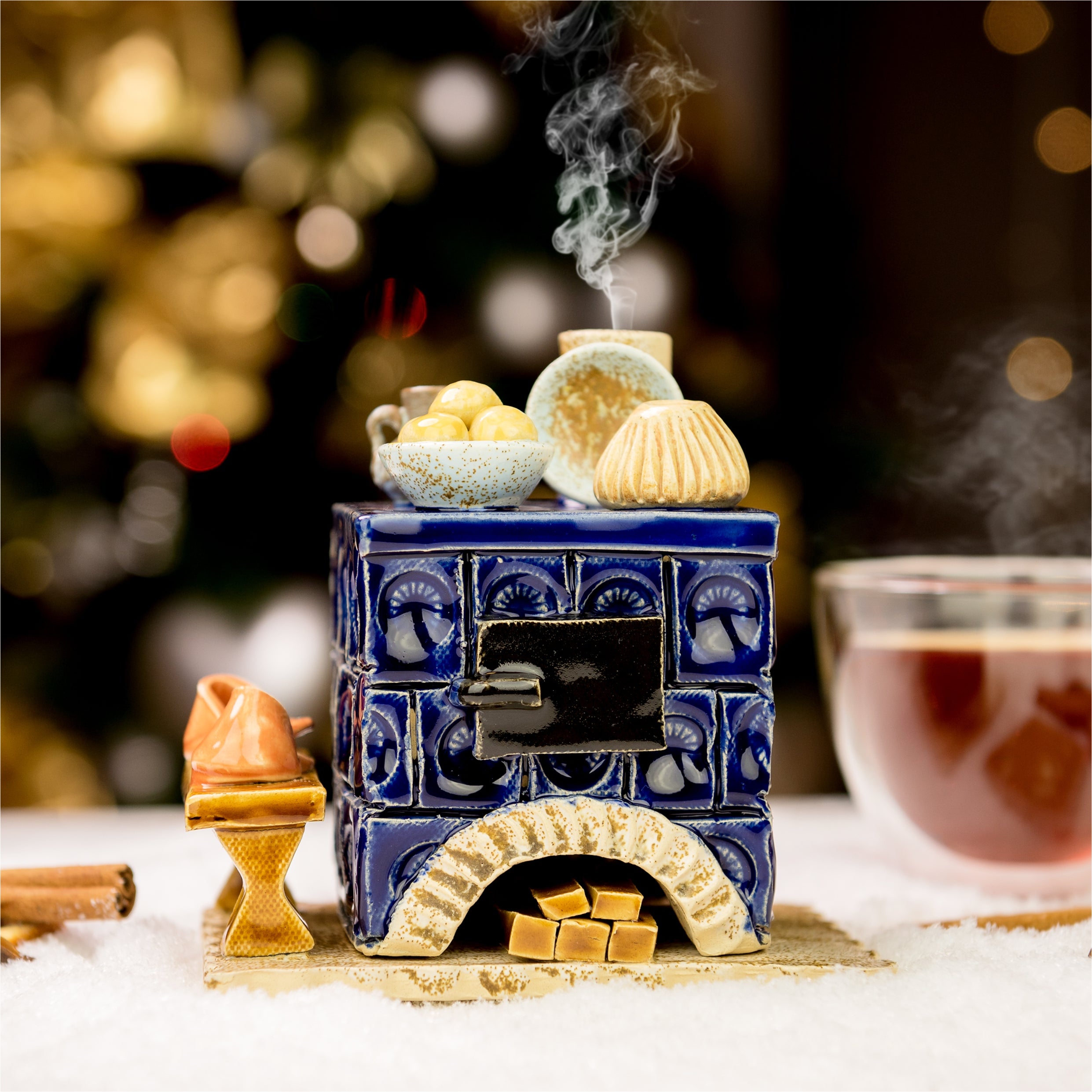 Der Keramik-Räucherofen Mini eckig, blau von Keramikwerkstatt Carola Busch ist ein handgefertigter Keramikofen, der auf Schnee steht und mit Essen dekoriert ist. Dampf steigt aus einem Topf auf, während Tee und Gewürze in der Nähe stehen. Verschwommene goldene Bokeh-Lichter im Hintergrund schaffen eine warme, festliche Atmosphäre.