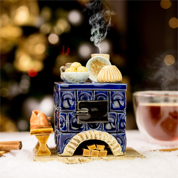 Der Keramik-Räucherofen Mini eckig, blau von Keramikwerkstatt Carola Busch ist ein handgefertigter Keramikofen, der auf Schnee steht und mit Essen dekoriert ist. Dampf steigt aus einem Topf auf, während Tee und Gewürze in der Nähe stehen. Verschwommene goldene Bokeh-Lichter im Hintergrund schaffen eine warme, festliche Atmosphäre.