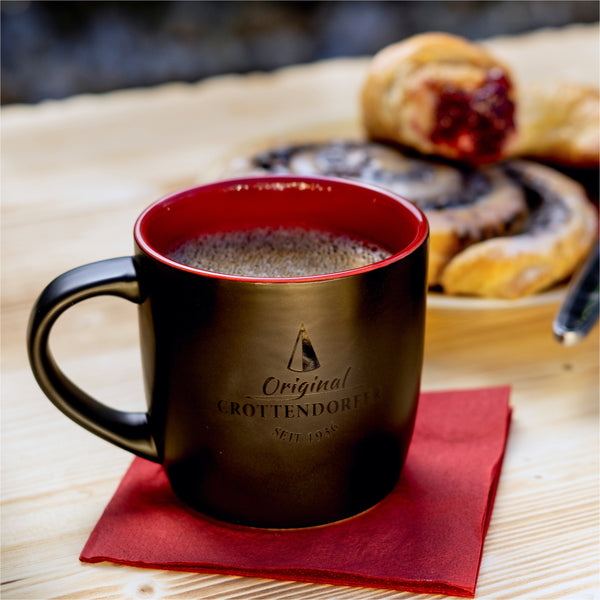 Eine mattschwarze 300 ml Keramiktasse von Original Crottendorfer Räucherkerzen mit roter Innenseite steht auf einer roten Serviette auf dem Holztisch. Im Hintergrund sind Gebäckstücke zu sehen, darunter eines mit roter Marmelade. Die Tasse ist spülmaschinengeeignet und verbindet Funktionalität mit Stil.