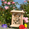 Eine hölzerne Erzgebirgslaterne mit Sonnenblumenschnitzerei ruht auf einer Steinoberfläche neben den Tischlaternen Sommerblumen der Drechslerei Kuhnert in Lila, Rot und Gelb. Die Szene zeigt rosa Blumen und üppiges grünes Blattwerk unter einem blauen Himmel und unterstreicht die traditionelle Handarbeit.