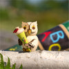 Der Mini Eule Schulanfänger von Drechslerei Kuhnert ist eine Holzfigur, die ein buntes Zapfengeschenk auf einem Birkenstamm aus nachhaltig gewonnenem Holz hält. Große ABC-Buchstaben schmücken den Hintergrund vor leuchtendem Grün und heben Kuhnerts Handwerkskunst hervor.
