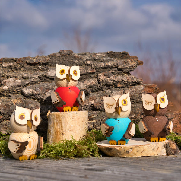 Vier MINI Eulen der Drechslerei Kuhnert, genannt Mini Eule natur, in Handarbeit gefertigt, tragen rote, blaue und braune Outfits und sitzen auf Baumstammscheiben vor einem Hintergrund aus Rinde auf einer moosbedeckten Fläche unter einem klaren blauen Himmel.