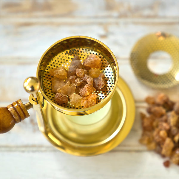 Auf einer hellen Holzoberfläche steht ein goldenes Räuchergefäß mit Holzgriff, gefüllt mit Räucherharz Myrrhe 20g von Original Crottendorfer Räucherkerzen. Zusätzliche Myrrhe- und Räucherstoffe sind in der Nähe verstreut, ideal für die Schaffung einer Schutz- und Reinigungsweihrauch-Atmosphäre.
