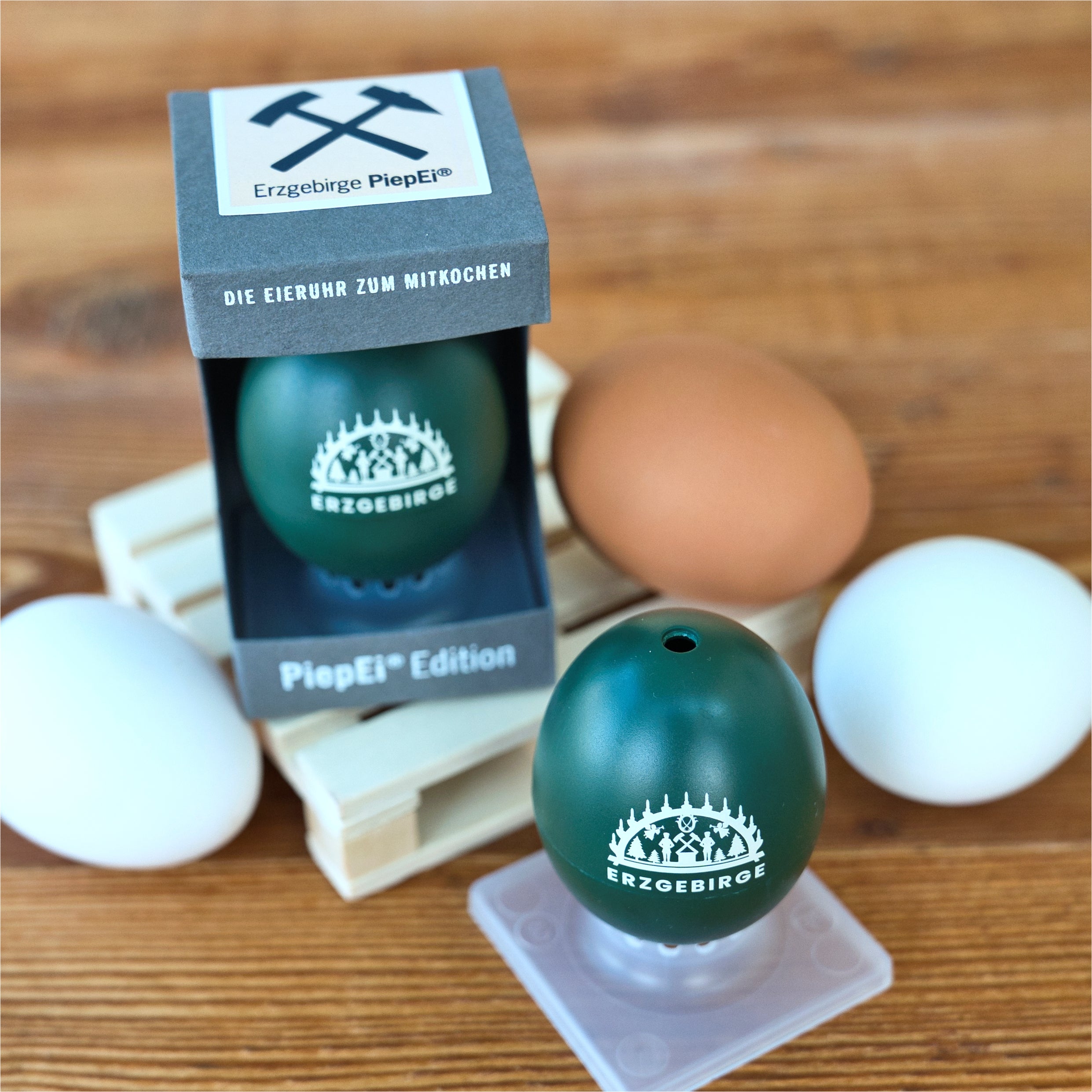 Das BrainStream PiepEi „Erzgebirge“, edel präsentiert auf einer Holzpalette in der Verpackung mit Bergbau-Emblem, ist umgeben von drei weißen und einem braunen Ei und verspricht perfekt gegarte Eier auf charmanter Holzoberfläche.