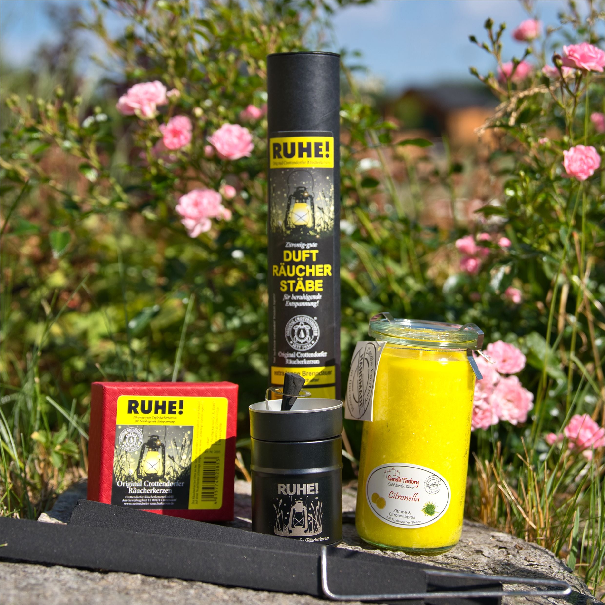 Eine Sammlung aromatischer Produkte wird elegant im Freien inmitten von rosa Blumen und üppigem Grün präsentiert. Mit der Duftkerze/Windlicht „Citronella“ Mini Jumbo-Kerze von Candle Factory in einem Glas, Räucherstäbchen in einer Tube und vielem mehr strahlt diese Szene die Ruhe der Natur auf der Steinoberfläche aus.