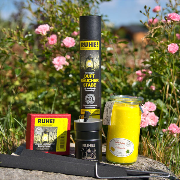 Eine Sammlung aromatischer Produkte wird elegant im Freien inmitten von rosa Blumen und üppigem Grün präsentiert. Mit der Duftkerze/Windlicht „Citronella“ Mini Jumbo-Kerze von Candle Factory in einem Glas, Räucherstäbchen in einer Tube und vielem mehr strahlt diese Szene die Ruhe der Natur auf der Steinoberfläche aus.
