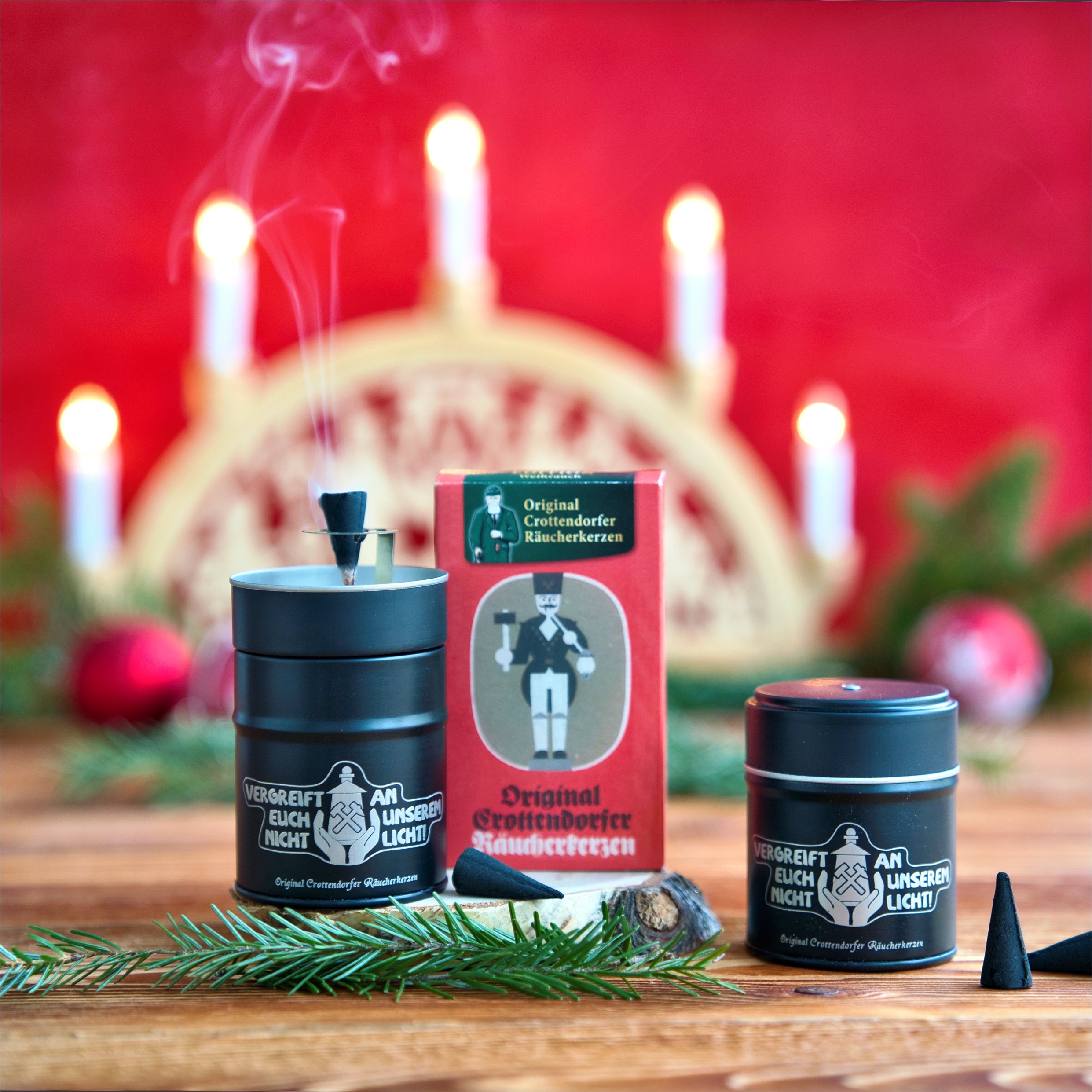 Auf Holz mit Tannenzweigen sind zwei schwarze Dosen und eine rote Schachtel Original Crottendorfer Räucherkerzen Räucherstäbchen ausgestellt. Ein „Vergreift euch nicht an unserem Licht“-Räucherfass, ein Schwibbogen und rote Ornamente schaffen eine festliche Szene, während brennendes Räucherwerk sanften Rauch für verstärktes Räuchervergnügen ausstößt.