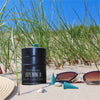 Eine schwarze Räucherfass-Sprüche von Original Crottendorfer Räucherkerzen liegt auf Sandgras am Strand. Muscheln, Sonnenbrillen, ein geflochtener Hut und kegelförmige Räucherstäbchen evozieren eine Atmosphäre des Räuchervergnügens. Der strahlend blaue Himmel lässt auf einen sonnigen Tag schließen.