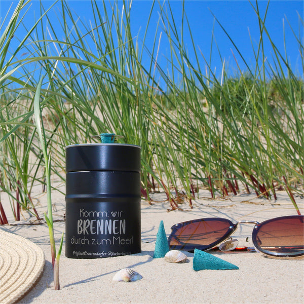 Eine schwarze Räucherfass-Sprüche von Original Crottendorfer Räucherkerzen liegt auf Sandgras am Strand. Muscheln, Sonnenbrillen, ein geflochtener Hut und kegelförmige Räucherstäbchen evozieren eine Atmosphäre des Räuchervergnügens. Der strahlend blaue Himmel lässt auf einen sonnigen Tag schließen.