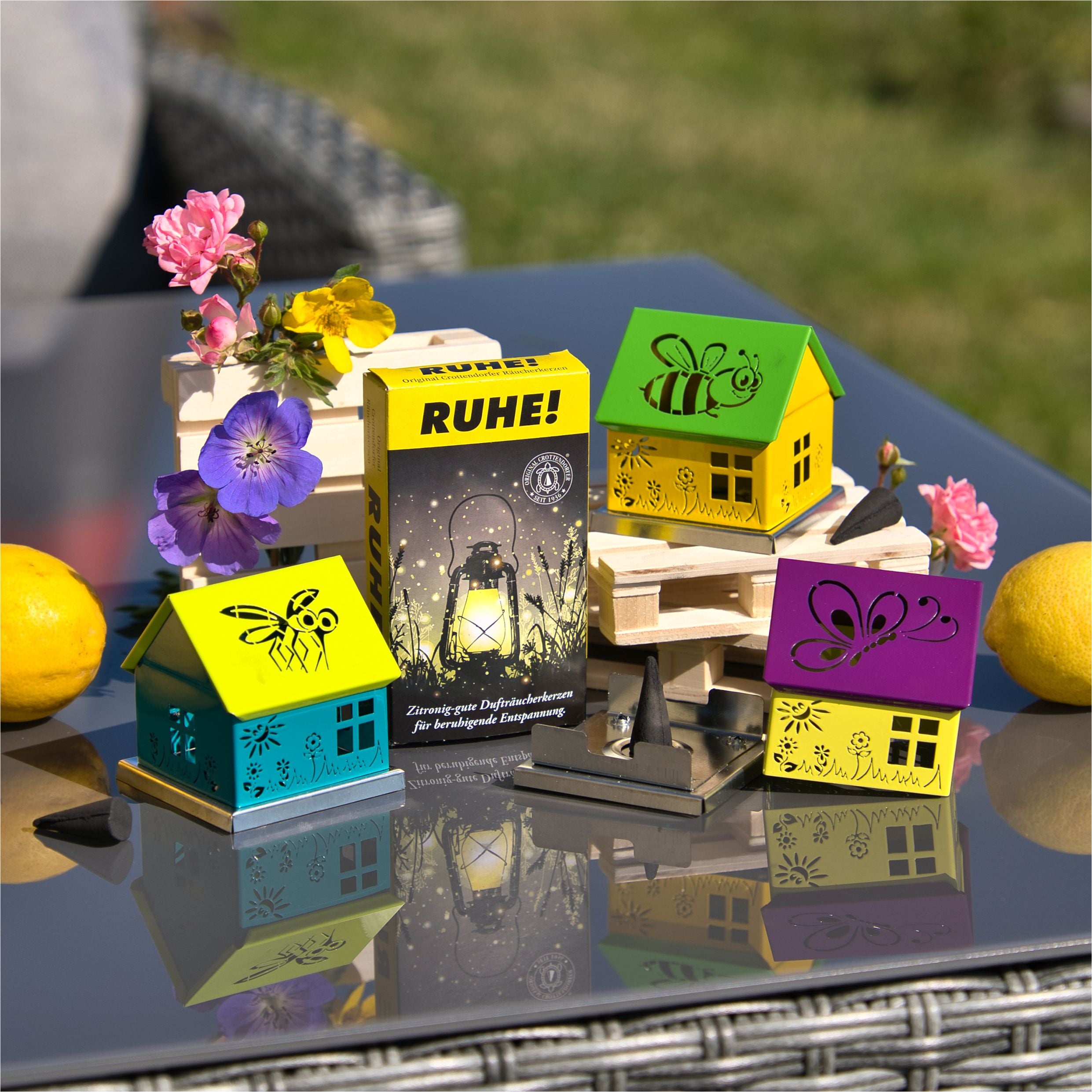 Die „Räucherhäuschen Biene grün/gelb“ der Schürer GmbH sind farbenfrohe Teelichthalter in Häuschenform mit Blumen- und Insektenausstanzungen. Ein „RUHE!“-Windlichtkasten, Blumen im Glas und frische Zitronen komplettieren diese bezaubernde Outdoor-Dekosidee auf einem Glastisch.