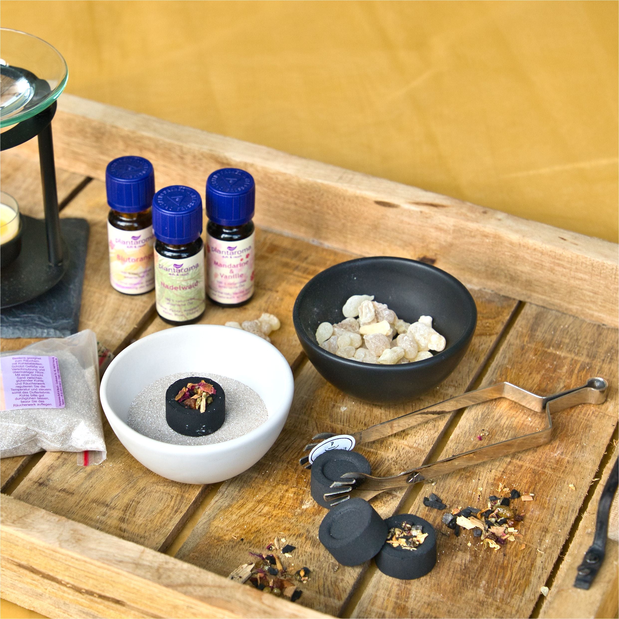 Auf einem Holztablett ist ein heiteres Aromatherapie-Setup zu sehen: eine schwarze Schale mit Harzstücken, eine weiße Schale mit Sand und schnell entzündliche Räucherkohle von Original Crottendorfer Räucherkerzen. Flaschen mit ätherischen Ölen, Metallzangen und ein Teelichthalter bereichern das Ambiente auf einem gelben Hintergrund.