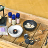Auf einem Holztablett ist ein heiteres Aromatherapie-Setup zu sehen: eine schwarze Schale mit Harzstücken, eine weiße Schale mit Sand und schnell entzündliche Räucherkohle von Original Crottendorfer Räucherkerzen. Flaschen mit ätherischen Ölen, Metallzangen und ein Teelichthalter bereichern das Ambiente auf einem gelben Hintergrund.