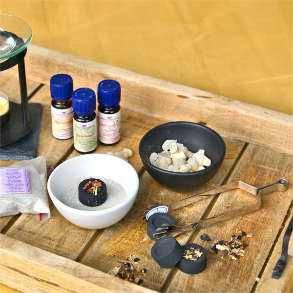 Auf einem Holztablett ist ein heiteres Aromatherapie-Setup zu sehen: eine schwarze Schale mit Harzstücken, eine weiße Schale mit Sand und schnell entzündliche Räucherkohle von Original Crottendorfer Räucherkerzen. Flaschen mit ätherischen Ölen, Metallzangen und ein Teelichthalter bereichern das Ambiente auf einem gelben Hintergrund.