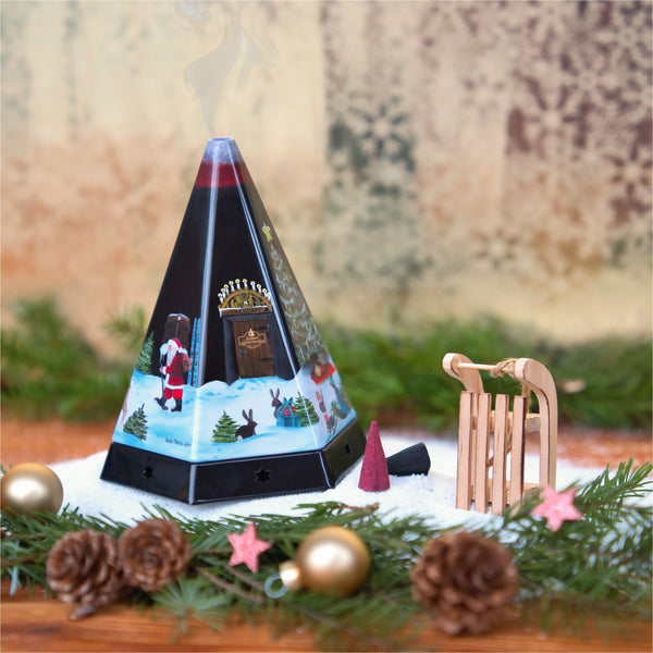 Die Räucherpyramide aus Metall von Original Crottendorfer Räucherkerzen strömt Rauch aus einem festlichen Räuchergefäß, umgeben von einem Holzschlitten, Tannenzweigen und Tannenzapfen auf einer schneeähnlichen Oberfläche. Die Metallpyramide zeigt weihnachtliche Motive, darunter Weihnachtsmann und Rentier.