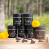 Fünf schwarze Original Crottendorfer Räucherkerzen Räucherfass Sprüche-Gläser sind pyramidenförmig auf Holz gestapelt und erwecken den Charme eines traditionellen Räucherkerzenhalters. Gelbe Blumen und Räucherkegel bereichern die Szene vor einem verschwommenen Hintergrund im Freien mit üppigem Grün und Bäumen.