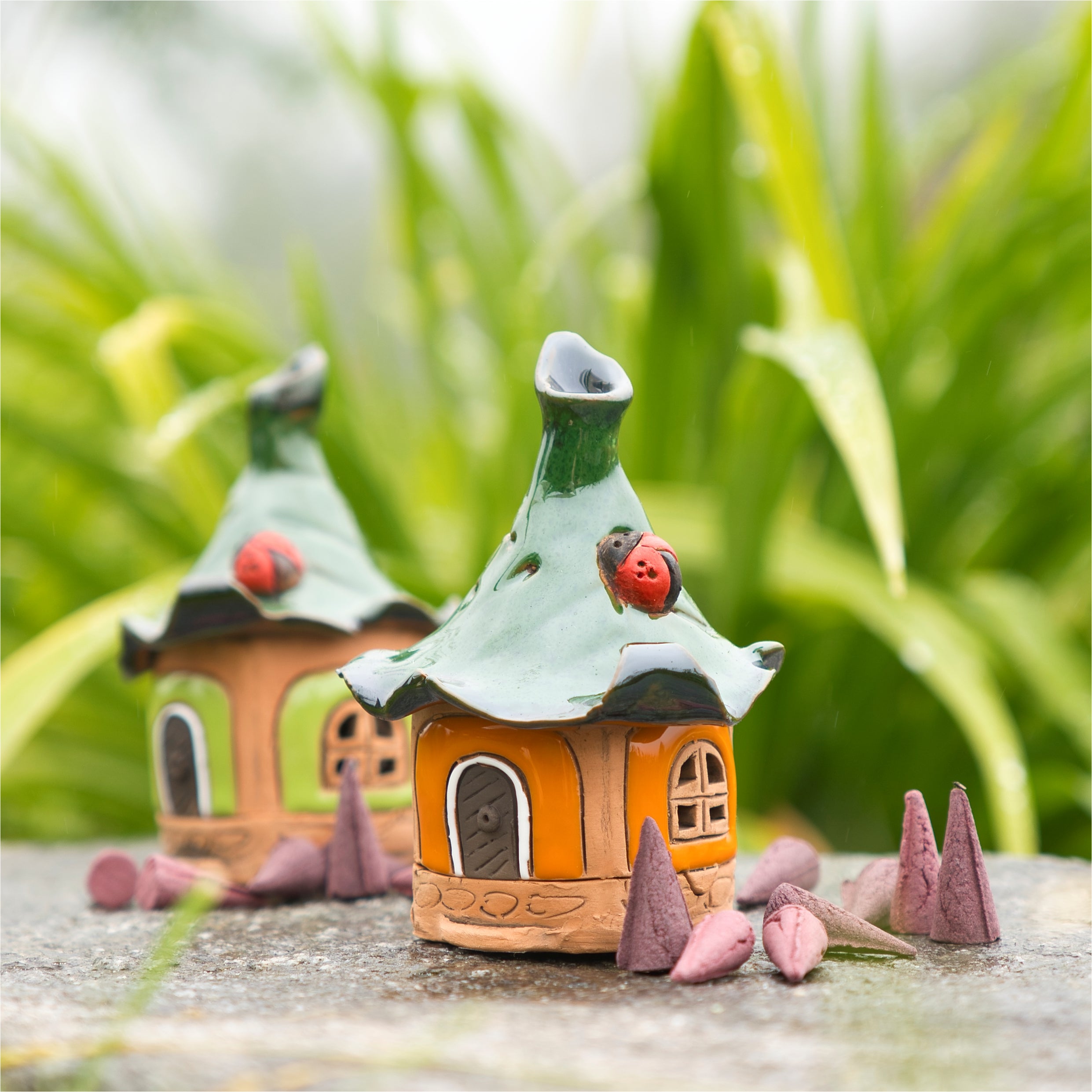 Zwei skurrile orange Mini-Designs von Räucherhaus Keramik mit aufgemalten Marienkäfern von Original Crottendorfer sitzen zwischen grünen Pflanzen auf einer Steinoberfläche, umgeben von kegelförmigen Räucherstäbchen, und erzeugen so einen zauberhaften Märchengarten-Effekt.