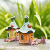Zwei skurrile orange Mini-Designs von Räucherhaus Keramik mit aufgemalten Marienkäfern von Original Crottendorfer sitzen zwischen grünen Pflanzen auf einer Steinoberfläche, umgeben von kegelförmigen Räucherstäbchen, und erzeugen so einen zauberhaften Märchengarten-Effekt.