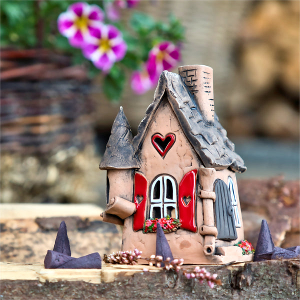 Das Räucherhaus Keramik mit Herz von Original Crottendorfer ist ein skurriles Miniatur-Tonhaus mit grauem Dach und roten Herzakzenten, eingebettet zwischen lila Blütenblättern und Grünpflanzen. Vor dem Hintergrund verschwommener lila Blumen in einer rustikalen Szenerie ist dieses charmante Stück in limitierter Auflage erhältlich.