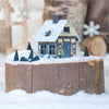 Das Räucherhäuschen Winterfreude von Original Crottendorfer Räucherkerzen ist ein bezauberndes, schneebedecktes Miniaturhaus mit illustrierten Fenstern und einem Schornstein auf einem Holzstumpf. Umgeben von immergrünen Bäumen fängt es eine gemütliche Winterlandschaft mit verschwommenen Schneeflocken und Baumstämmen im Hintergrund ein.