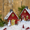 Zwei kleine Räucherhäuschen Wintermotiv von Original Crottendorfer Räucherkerzen, mit verschneiten Winterlandschaften, festlichen Dekorationen und Schneemännern, ruhen auf Kunstschnee. Warme Lichter leuchten sanft im Hintergrund, während ein metallener Räucherkerzenhalter dieses gemütliche Winterwunderland noch verstärkt.