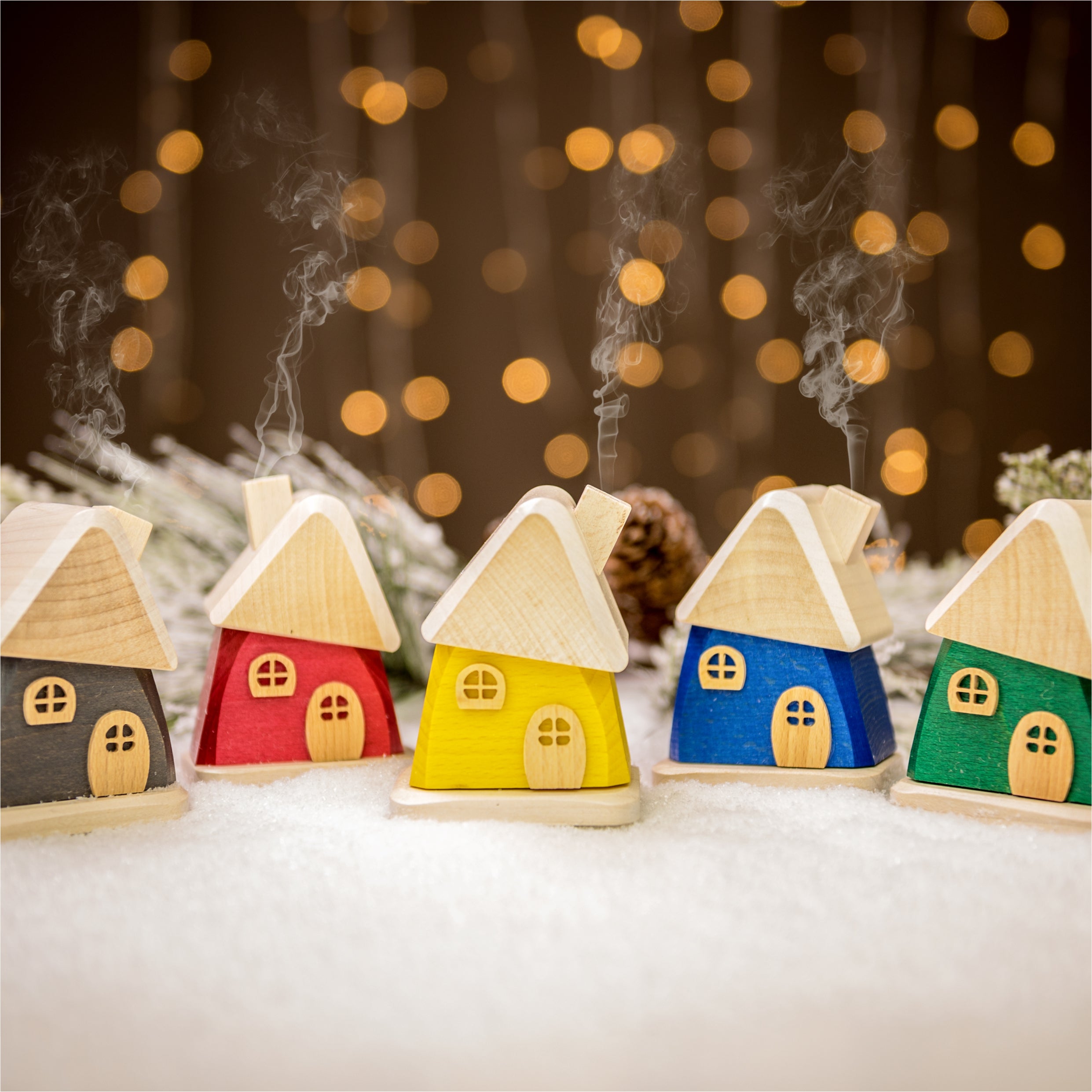 Fünf farbenfrohe, handgefertigte Räucherhäuschen aus Holz von Holzwaren Egermann (rot, gelb, blau, grau, grün) stehen auf einer schneebedeckten Oberfläche mit sanftem Bokeh-Licht im Hintergrund. Aus den Schornsteinen dieser Häuser im Erzgebirge-Stil steigt sanft Rauch auf und schafft eine warme, festliche Atmosphäre.