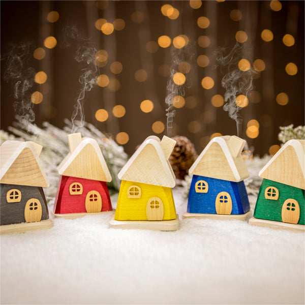 Fünf farbenfrohe, handgefertigte Räucherhäuschen aus Holz von Holzwaren Egermann (rot, gelb, blau, grau, grün) stehen auf einer schneebedeckten Oberfläche mit sanftem Bokeh-Licht im Hintergrund. Aus den Schornsteinen dieser Häuser im Erzgebirge-Stil steigt sanft Rauch auf und schafft eine warme, festliche Atmosphäre.