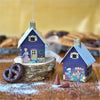 Zwei kleine, dekorierte Blechhäuschen in Lebkuchenform stehen auf einer Holzfläche, eines davon auf einem Baumstumpf neben Brezeln und Zuckertüten. Die metallischen Ornamente sind mit Menschen- und Tiermotiven verziert und passen als Räucherhäuschen Weihnachtsbäckerei von Original Crottendorfer Räucherkerzen perfekt in jede festliche Umgebung.