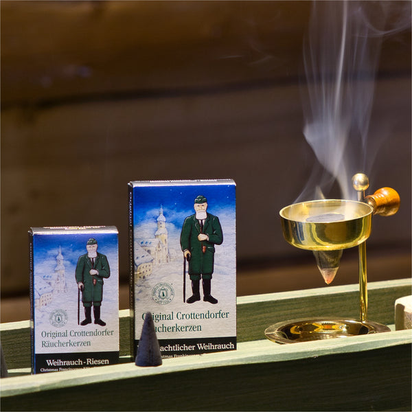 Zwei Schachteln mit Original Crottendorfer Räucherkerzen stehen neben einem Weihrauchbrenner aus Messing. Das Display enthält eine detaillierte Räucheranleitung und zeigt eine stehende Figur vor einer verschneiten Kulisse.