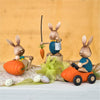 Drei Stupsi Hase Möhrenangler-Osterhasen aus Holz von Drechslerei Kuhnert in farbenfroher Kleidung sind ausgestellt. Handarbeitsdetails zeigen einen mit einer Karotte auf einem Stock, einen anderen auf einer großen Karotte und den dritten, der ein Karottenauto fährt. Geschmückt mit gesprenkelten Eiern und grünem Band auf orangefarbenem Hintergrund.
