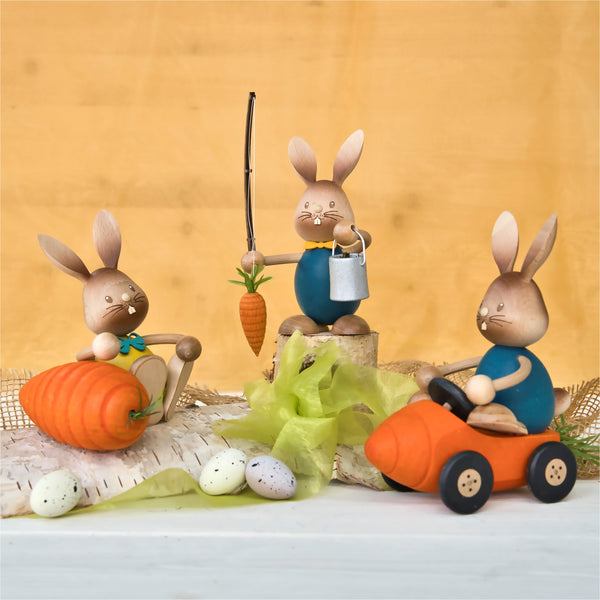 Drei Stupsi Hase Möhrenangler-Osterhasen aus Holz von Drechslerei Kuhnert in farbenfroher Kleidung sind ausgestellt. Handarbeitsdetails zeigen einen mit einer Karotte auf einem Stock, einen anderen auf einer großen Karotte und den dritten, der ein Karottenauto fährt. Geschmückt mit gesprenkelten Eiern und grünem Band auf orangefarbenem Hintergrund.