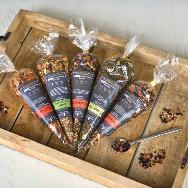 Fünf kegelförmige Beutel Original Crottendorfer Grünteemischung "Bergmanns-Glück" mit bunten Etiketten sind auf einem Holztablett angeordnet. Ein Teelöffel mit losen Teeblättern liegt daneben auf einer hellen Unterlage.