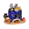 Der Keramik-Räucherofen, Mini rund, blau der Keramikwerkstatt Carola Busch zeigt einen blauen Kachelofen mit Töpfen und Geschirr darauf, eine schwarze Katzenfigur daneben, Holzscheite darunter und ein kleines geschwungenes rotes Dekor, das an die Handwerkskunst des Erzgebirges erinnert.