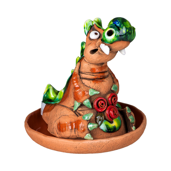 Der Original Crottendorfer Räucherdrache hat grüne Zacken, ausdrucksstarke Augen, ein verspieltes Lächeln und sitzt in einem runden braunen Untersetzer mit roten Rosen auf der Brust.